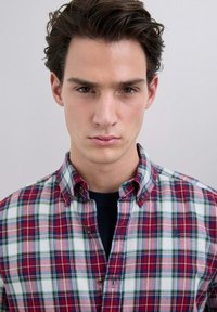 Giovane uomo con capelli scuri che indossa una camicia a quadri rossa, bianca e blu sopra una maglia nera, rivolto verso l'osservatore con un'espressione neutra.