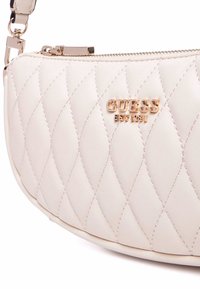 Borsa a tracolla trapuntata color crema con finitura lucida, caratterizzata da un logo "GUESS" in tonalità oro e chiusura con cerniera. Inclusa tracolla regolabile.
