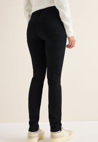 Höga svarta jeans i denim, med slim fit, två bakfickor, och matchade med ljusa sneakers mot en neutral bakgrund.