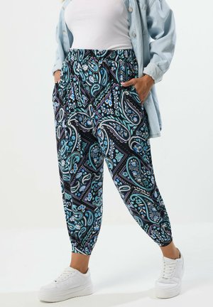 Kobieta ubrana w luźne spodnie w kolorowy wzór paisley w odcieniach niebieskiego i fioletu, białe trampki, biały top oraz jasną kurtkę dżinsową, z rękami w kieszeniach.