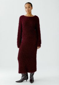 Burgunderfarbiges, flauschiges Strickkleid mit langen Ärmeln und einem runden Halsausschnitt, das bis zur Mitte des Wadenbereichs reicht; kombiniert mit spitzen, schwarzen Stiefeletten.
