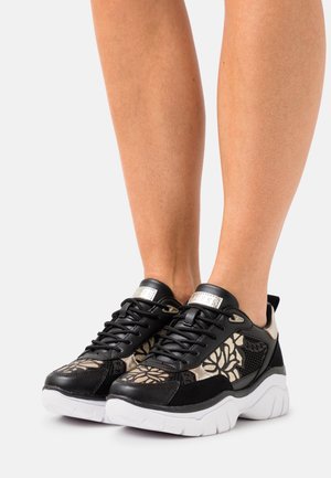 Czarno-złote sneakersy w kwiatowy wzór z grubymi białymi podeszwami, noszone na stopach, przedstawione na jednolitym jasnym tle.