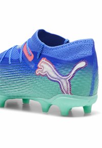 Cramponi de fotbal sintetici albastre și verzi, cu o suprafață texturată, accente roz și un logo Puma alb proeminent pe lateral.