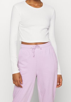 Personne portant un crop top blanc côtelé à manches longues et un pantalon de survêtement violet clair avec cordon de serrage sur un fond uni.