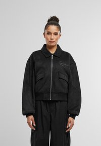 Jachetă neagră scurtă dintr-un material neted, cu fermoar frontal, guler mare și două buzunare frontale cu detalii subtile ale logo-ului.