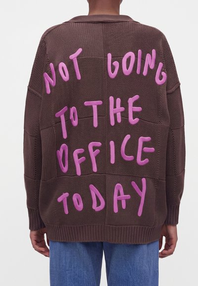 Personne portant un pull en tricot marron avec des lettres roses indiquant "NOT GOING TO THE OFFICE TODAY" dans le dos.