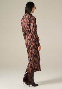 Robe à manches longues en tissu satiné, ornée d'un motif géométrique brun et sombre, avec une taille cintrée et une longueur en dessous des genoux. Assortie à des bottes hautes.