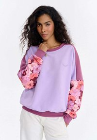 Lilla dressipluus roosade ja bordoopunaste lillemustriliste varrukatega, millel on rinnal lihtne logo ning soonikkoes kaelus ja mansetid.