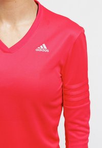 Rød atletisk top med v-hals, lange ærmer med vandrette striber og et hvidt Adidas-logo på brystet.