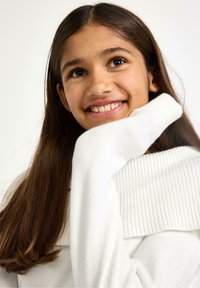 Maglione bianco a maglia con collo alto a coste, texture morbida e maniche lunghe, indossato da una persona con capelli lunghi e scuri.