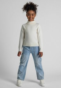 Cremfarbener Rollkragenpullover kombiniert mit hellblauen Cargo-Hosen. Das Outfit besticht durch einen strukturierten Stoff und enganliegende Bündchen, ergänzt durch weiße Sneakers.