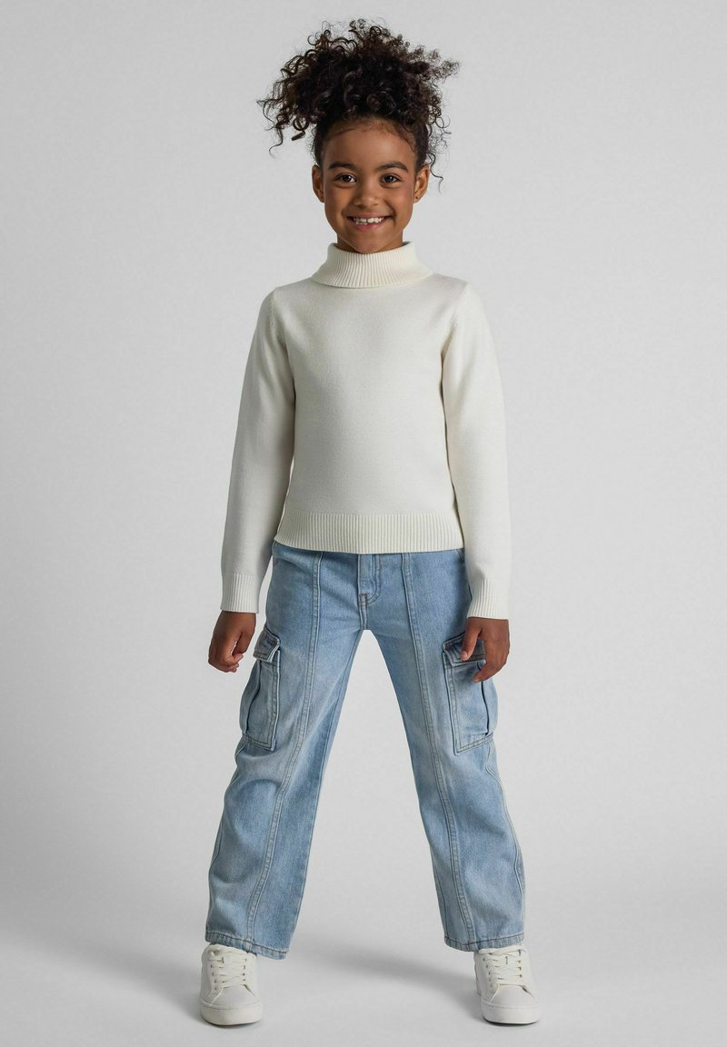 Cremfarbener Rollkragenpullover kombiniert mit hellblauen Cargo-Hosen. Das Outfit besticht durch einen strukturierten Stoff und enganliegende Bündchen, ergänzt durch weiße Sneakers.