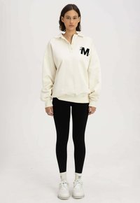 Crèmekleurige sweatshirt met een kraag en geborduurd 'M'-detail, gecombineerd met zwarte leggings en witte sneakers.