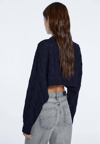 Pull en maille cropped bleu marine avec motif torsadé texturé et manches larges, porté avec un jean gris clair taille haute.