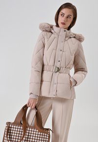 Vrouw in een beige gewatteerde jas met een bontcapuchon en riem, die een bruin-witte geblokte handtas vasthoudt, terwijl ze zijwaarts kijkt tegen een effen achtergrond.