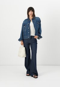 Denimjakke, blå, med en cropped design, store lommer og sølvknapper. Kombinert med flared denimbukser og en hvit teksturert håndveske.