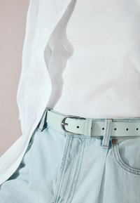 Ceinture en cuir vert menthe avec une boucle en D en métal argenté, portée sur un jean bleu clair, associée à un débardeur blanc à côtes et une chemise à boutons.