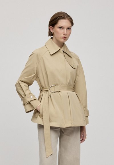 Trench-coat beige ceinturé avec un grand col, des accents de boutons et des manches tapered. Porté sur un pantalon clair, avec une coupe structurée.