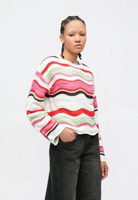 Νεαρός με πλεγμένα μαλλιά, που φοράει πολύχρωμο ριγέ πλεκτό πουλόβερ και μαύρο τζιν, στέκεται με το ένα χέρι στ ην τσέπη μπροστά σε λευκό φόντο.