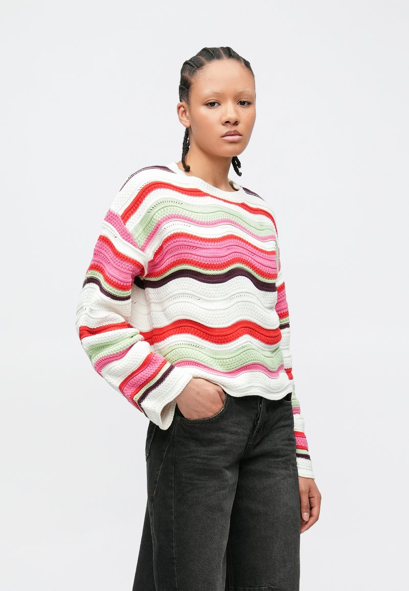 Νεαρός με πλεγμένα μαλλιά, που φοράει πολύχρωμο ριγέ πλεκτό πουλόβερ και μαύρο τζιν, στέκεται με το ένα χέρι στην τσέπη μπροστά σε λευκό φόντο.