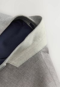 Gros plan d'un col de blazer gris montrant l'extérieur en tissu texturé et la doublure intérieure lisse et foncée avec des finitions de couture soignées.