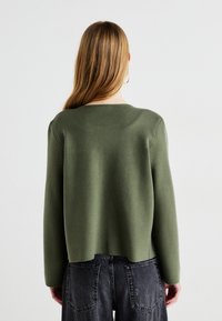 Πράσινο, μακρυμάνικο πουλόβερ με στρογγυλή λαιμόκοψη, φτιαγμένο από μαλακό ύφασμα, με ελαφρώς κοντή σχεδίαση, συνδυασμένο με ψηλόμεσα μαύρα τζιν.