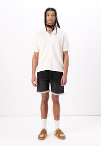 Chemise beige clair à manches courtes, short en denim noir avec des ourlets retroussés, chaussettes blanches et baskets marron. Le modèle se tient devant un fond blanc.