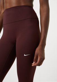 Bordeaux høytlivs leggings laget av elastisk materiale, med en hvit Nike-logo på venstre lår og glatt, strukturert stoff.