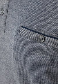Nahaufnahme eines strukturierten Poloshirts aus blauem und weißem Stoff mit einer geknöpften Brusttasche, die einen kleinen dunklen Knopf und detaillierte Nähte aufweist.