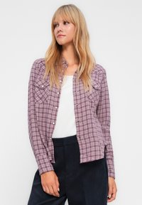 Chemise à carreaux violette et blanche, avec un devant à boutons, deux poches poitrine et des manches longues, associée à un pantalon sombre.