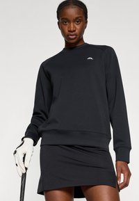 Fekete pulóver sima, rugalmas anyagból, kerek nyakkivágással és bordázott mandzsettákkal. Fekete szoknyával párosítva, amelynek finom esése van.