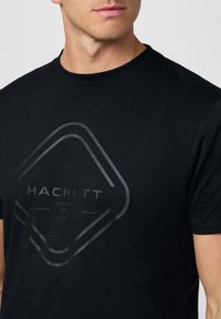 Camiseta de algodón negra con un gran gráfico en forma de diamante brillante que incluye la palabra "HACKETT" y texto adicional en un sutil tono gris.