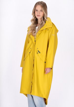 Fiatal nő, aki hosszú sárga esőkabátot visel elején zsebekkel és kapucnival, egyszínű fehér háttér előtt áll.