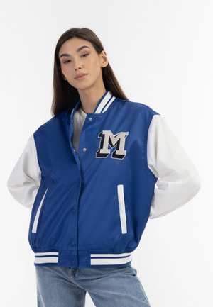 Młoda kobieta w niebiesko-białej kurtce varsity z dużą literą M na piersi, pozująca z rękami za plecami.