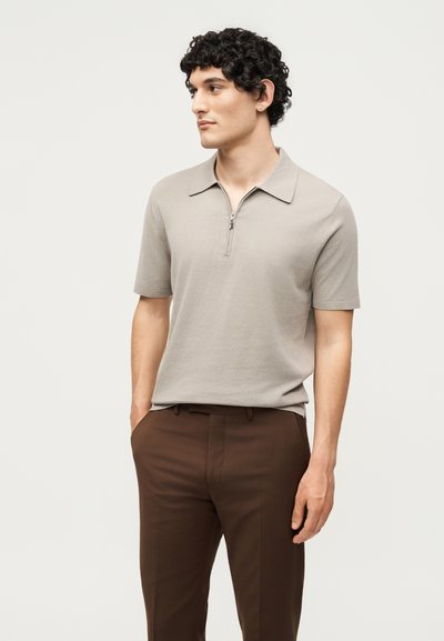 Άνδρας με σγουρά μαλλιά που φοράει μπεζ κοντομάνικη πλεκτή μπλούζα με φερμουάρ και καφέ παντελόνι, στέκεται με τα χέρια στις τσέπες, κοιτάζοντας προς τα αριστερά.