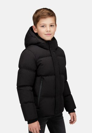 Jongen met lichtbruin haar die een zwarte gewatteerde jas met capuchon en donkere broek draagt, staand tegen een effen lichte achtergrond.