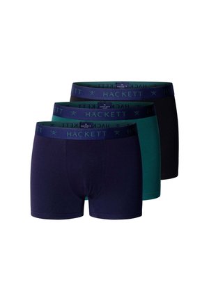 Jonge man met kort krullend haar staat op blote voeten tegen een effen muur en draagt donkerblauwe nauwsluitende boxershorts.