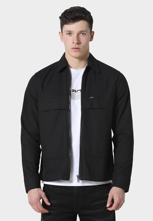 Ung mann med kort mørkt hår iført en svart glidelåsjakke over en hvit T-skjorte med grafisk trykk, stående foran en ensfarget lys bakgrunn.