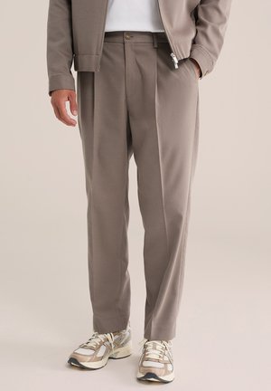 Homme portant un pantalon taupe sur mesure, des baskets beige et blanches, une veste taupe assortie et une chemise blanche sur un fond uni.