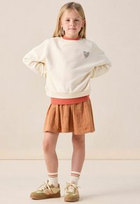 Sweatshirt crème avec des bords côtelés orange, patch en forme de cœur ; jupe texturée couleur rouille ; baskets vertes et beiges avec des chaussettes rayées blanches.