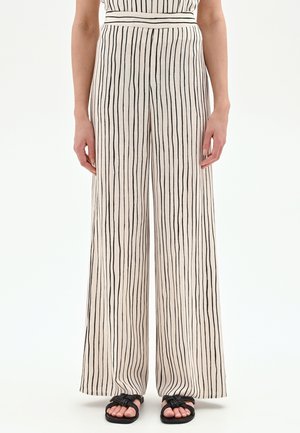 Pantalon beige à jambes larges avec des rayures verticales noires irrégulières, porté avec des sandales noires à lanières tressées, sur une personne debout.