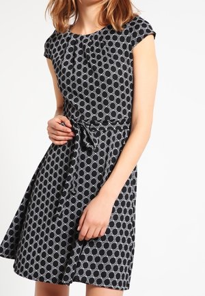 Nainen, joka pukeutunut mustaan mekkoon, jossa on valkoinen pyöreä kuvio, lyhyet hihat ja sidottu vyö vyötäröllä.