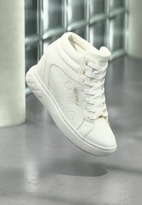 Baskets montantes blanches avec des motifs de logo en relief, des lacets plats, des accents dorés et une semelle texturée. Matériau synthétique lisse.