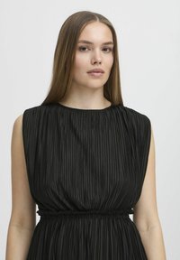 Abito nero plissettato con scollo rotondo e vita elastica. Il tessuto ha una texture liscia con pieghe verticali in tutto l'abito.