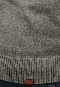 Pull en tricot gris avec un ourlet côtelé et une petite étiquette en cuir. Texture douce, épaisseur moyenne et subtiles variations de couleur.