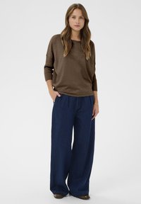 Braunes langärmliges Oberteil mit gerippten Bündchen, kombiniert mit weiten, dunkelblauen Jeans. Das Model steht mit den Händen in den Taschen vor einem neutralen Hintergrund.