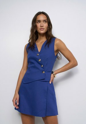 Mujer de pie contra un fondo liso, lleva un vestido azul sin mangas con botones dorados y falda, mano en la cadera, mirando ligeramente hacia un lado.