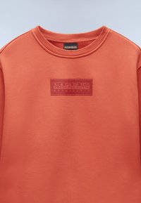 Sudadera color coral con cuello redondo, puños acanalados y un parche de logo texturizado en un coral más oscuro, hecha de una tela suave con un acabado liso.