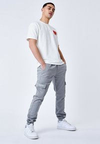 Camiseta blanca de manga corta con gráfico de corazón rojo, combinada con pantalones cargo grises claros que tienen bolsillos con cremallera y puños elásticos. Zapatillas blancas.