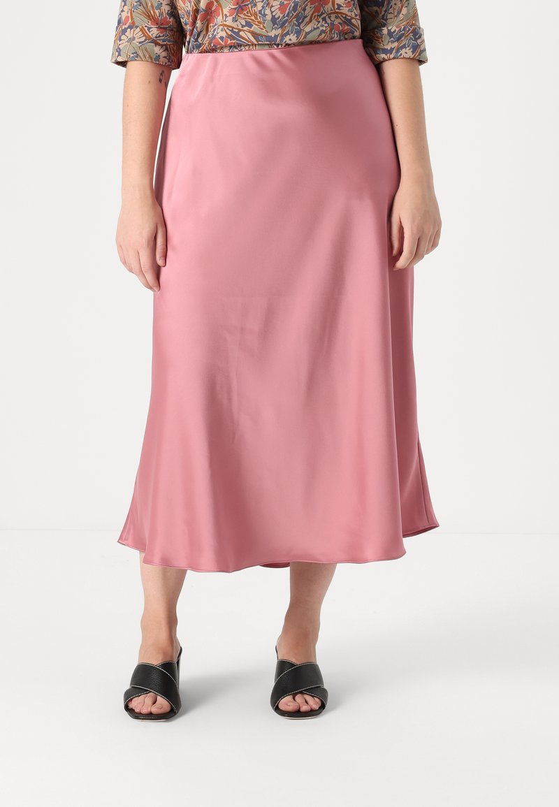 Jupe midi en satin rose clair avec une texture lisse et un subtil éclat, présentant une silhouette fluide et un ourlet droit. Portée avec des sandales à glissière noires.